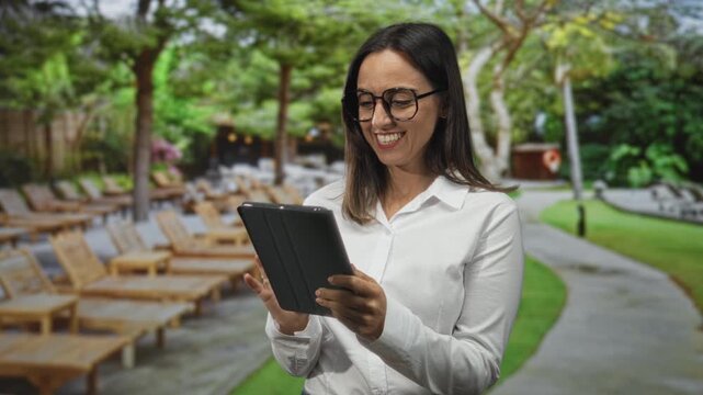 Woman holding tablet with both hands, smiling and tapping screen while standing beside wooden lounge chairs on a paved path near a building; joyful connection.