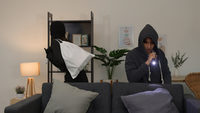 Masked and unmasked burglars sneaking behind a sofa in a living room
