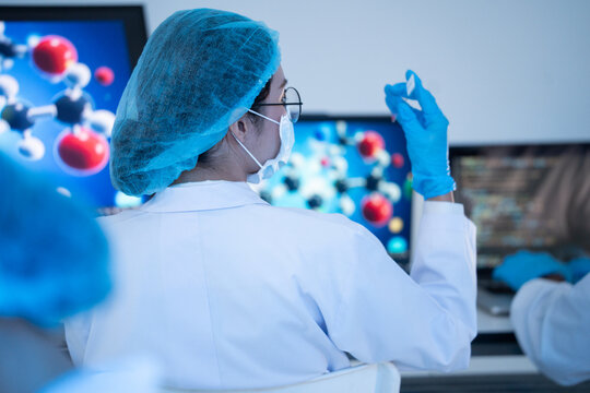 Back view of female scientist raising glass slide sample to examine clinical data in front of computer monitor showing molecule model inside medical laboratory