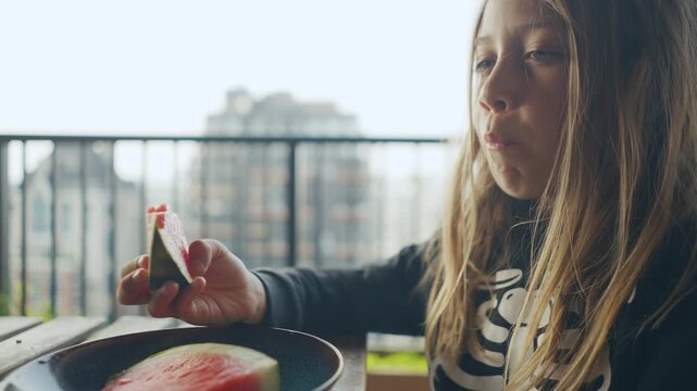 Blonde child eating watermelon on a sunny city balcony