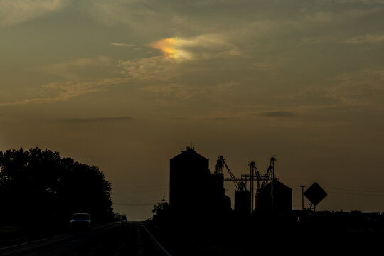 Darken silhouette of farm buildings and a road at dusk or dawn, with a sundog appearing overhead
