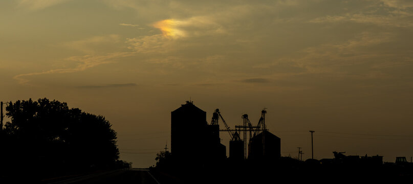 Darken silhouette of farm buildings and a road at dusk or dawn, with a sundog appearing overhead
