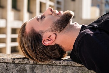 Side profile portrait of a young man resting on a concrete wall with modern architecture. © Valeria Vaskovats