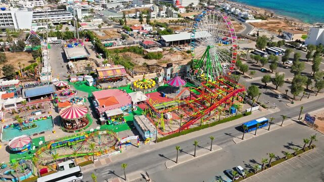 Drone view of Parko Paliatso amusement park in Ayia Napa, Cyprus. Bright rides, funfair attractions, nearby roads and the coastal town form a vibrant summer entertainment view.
