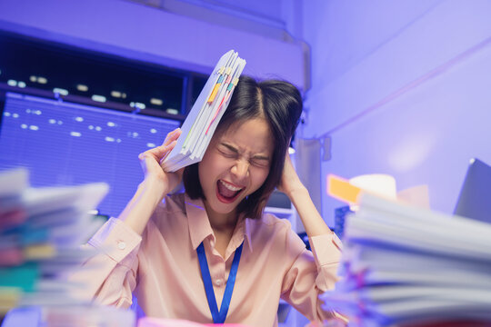 Stressed young Asian businesswoman overwhelmed with workload and holding documents while screaming in office.