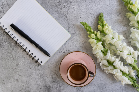 White flowers curl beside a coffee cup and notebook. Black pen rests on lined pages, ready for thought. Coffee steams gently, inviting quiet reflection. Concrete surface provides calm
