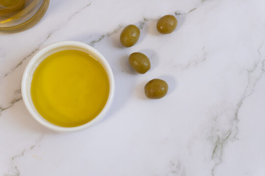 Fresh green olives beside golden olive oil in white bowl. Marble surface provides elegant, clean background. Natural, rustic feel evokes Mediterranean simplicity. Ideal for recipes, wellness