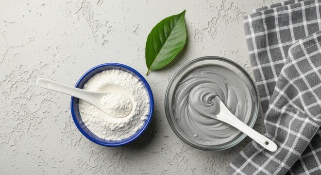 Top view of a blue bowl with white powder and a glass bowl with grey cosmetic clay mask