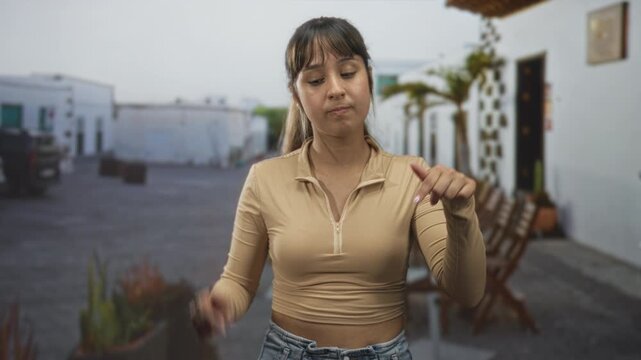 Young hispanic woman points finger down toward camera, bare midriff visible near white building entrance courtyard; dismissal.