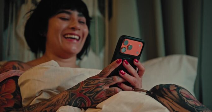 Young Tattooed Woman Laughing While Using Her Smartphone in Bed