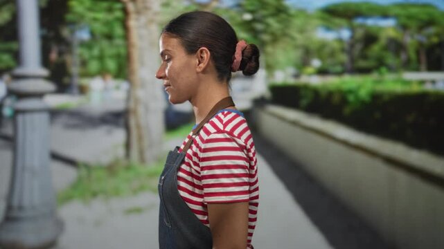 Woman pressing hands to cheeks and wincing beside a lamppost on a busy street wearing striped shirt and apron;distress.