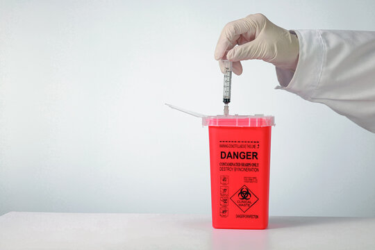 Cautious hand wearing glove performs clinical waste management by dropping sharp syringe needle into red plastic biohazard container