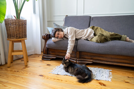 Relaxed woman comfortably lying on couch enjoying lazy day and time with domestic cat, stress free rest at home and procrastination, calm female zoomer patting pet on head while lounging on sofa.