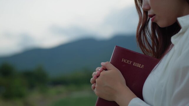 Faith concept: young Asian Christian woman embracing Holy Bible with eyes closed, praying outdoors by peaceful lake and mountain, symbolizing hope, peace and spirituality.
