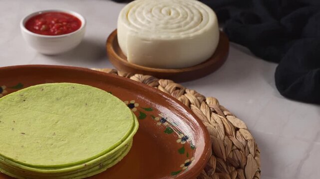 Green nopal corn tortillas stack with fresh Mexican cheese wheel and red salsa on clay plate