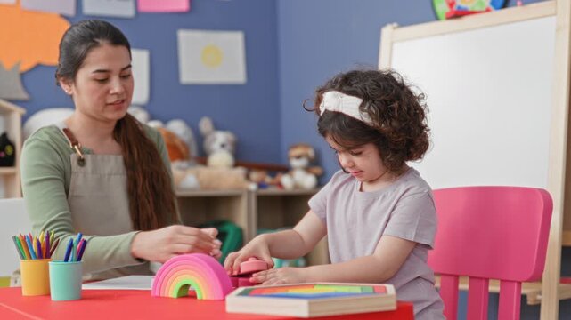 Woman teacher guiding child's hands on a rainbow stacking toy at a colorful classroom table with crayons and an easel in view; learning focus calm.
