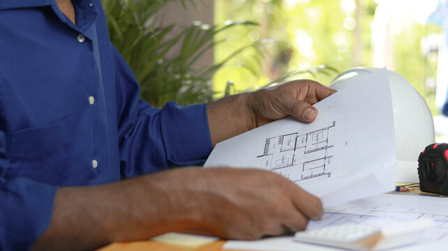 Professional engineer reviewing architectural blueprint and construction plans at a modern workspace with safety helmet. Concept of architecture, engineering, project management, and development.