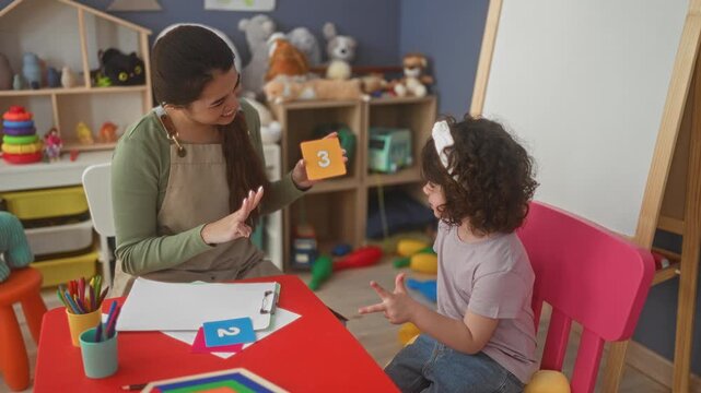 Teacher holds green flashcard showing 5 and raises palm while girl claps at a red table in building; learning concentration.