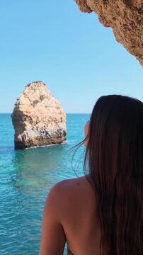 Back view of a young woman with long hair admiring the turquoise sea and rock formations on a sunny day. A beautiful summer holiday destination concept for travel and relaxation