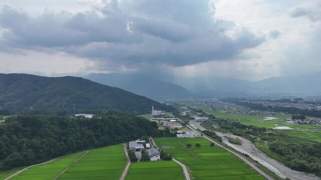 Aerial view shows riverbed between lush rice fields. Gravelly bed runs alongside green paddy rows. Trees frame the landscape, adding depth and texture. Village homes dot the distant horizon
