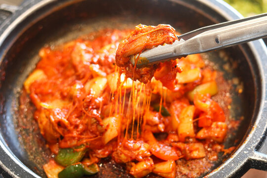 Lifting a piece of chicken from a Spicy Korean cheese buldak that's sizzling in a hot skillet topped with gooey melted cheese.