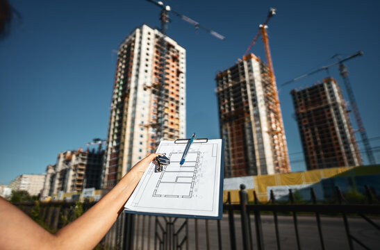 Real estate blueprint with house keys held in front of modern residential towers under construction. Concept of property investment, urban development, architecture planning and future home ownership.
