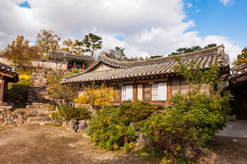 Fototapeta premium Captivating Exterior View of Traditional Korean Hanok House Featuring Historic Architecture, Tiled Roofs, and Autumn Garden Under Dramatic Blue Sky and Clouds