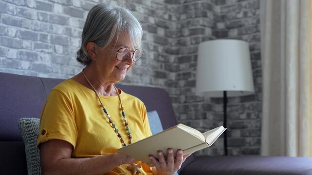 Happy older blonde woman sitting on comfortable sofa in living room, reading interesting book. Smiling elder retired grandmother enjoying leisure weekend time with favorite novel alone at home.