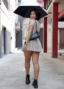 Asian woman in beige crop top and pleated skirt holding a black umbrella while walking outdoors in an alleyway