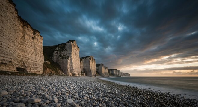 Storm clouds framing towering pale limestone cliffs above a lone pebble-strewn coastline, moody cinematic light
