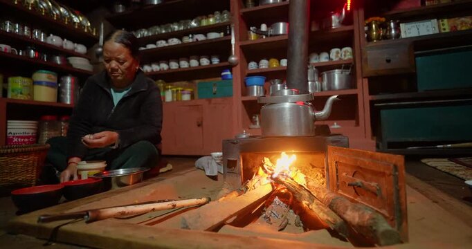 Annapurna Circuit, Manang, Nepal, camera shot of a local woman preparing food in her traditional firewood kitchen next to a burning wood stove with a metal kettle.