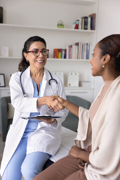 Cheerful confident mature doctor in lab coat shaking hands with young Black patient on consultation, expressing support. Young woman thanking practitioner for good healthcare news