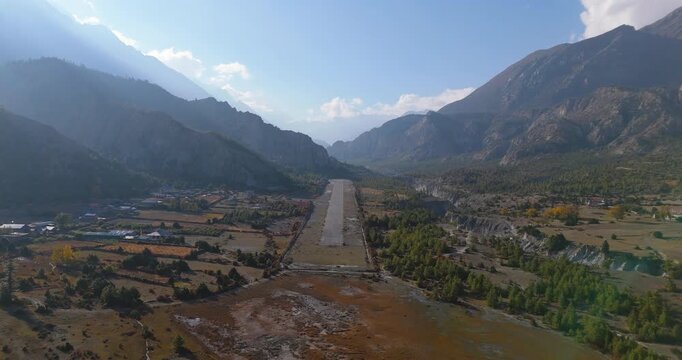 Annapurna Circuit, Manang Nepal, aerial gliding shot of the flowing Marsyangdi River running alongside the Humde Airport airstrip within a wide Himalayan valley.