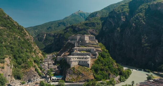 Aerial drone view of the historic Fort Bard in Italy's Aosta Valley featuring a winding river below and majestic Alpine peaks, showcasing breathtaking architecture and landscapes.