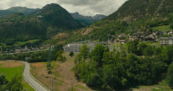 Drone flies above treetops toward an electric power substation nestled between mountains, showcasing high-voltage energy infrastructure in a scenic alpine landscape.