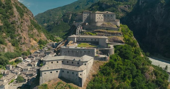Close-up drone orbit around the historic Fort Bard in Italy's Aosta Valley on a bright sunny day, highlighting its impressive fortress architecture and alpine surroundings.