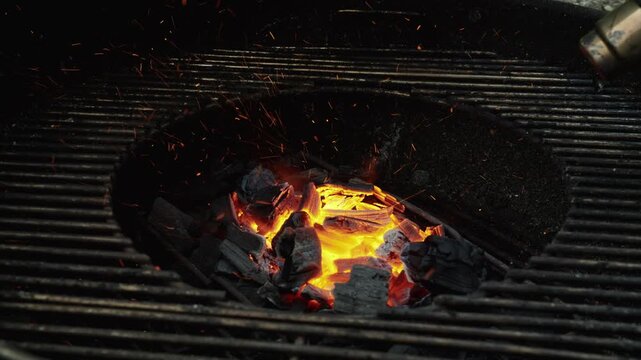 Hot charcoal briquettes burn in the center of a round backyard barbecue as orange embers and sparks rise through the dark metal grate.