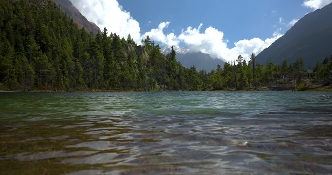 Annapurna Circuit, Manang, Nepal, camera shot of green lake water flowing gently in the foreground surrounded by dense pine forests and high Himalayan mountain peaks.