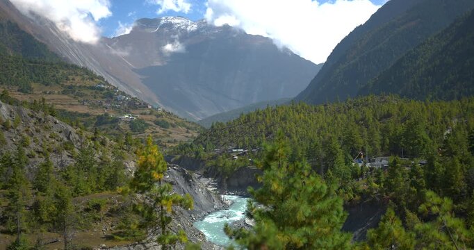 Annapurna Circuit, Manang, Nepal, wide camera shot of the Marsyangdi River flowing through a deep valley bordered by green pinewoods beneath high Himalayan mountain peaks.