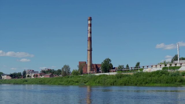 Tall red and white striped industrial factory chimney stands on green bank of Daugava river under clear blue sky in historic city of Daugavpils.