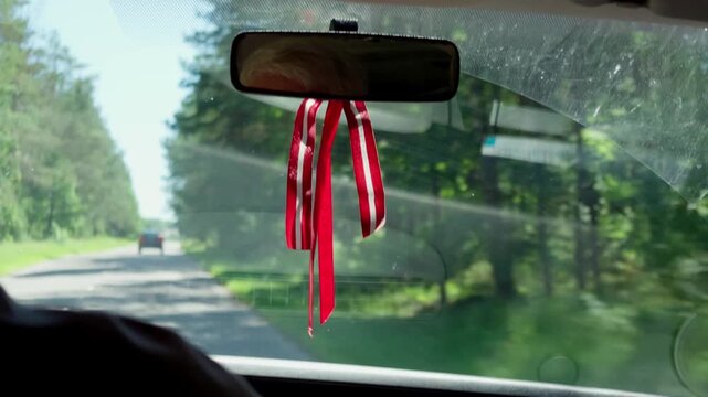 Red and white ribbon in traditional Latvian flag colors hangs on rearview mirror inside moving vehicle. Car travels down narrow asphalt road through dense green woods.