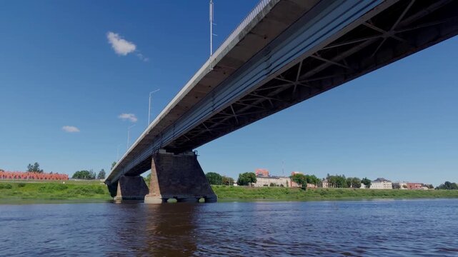 Camera passes slowly beneath concrete bridge span over calm river water during sunny summer day. Low angle shot captures massive pillar supports and steel girders under clear blue sky.