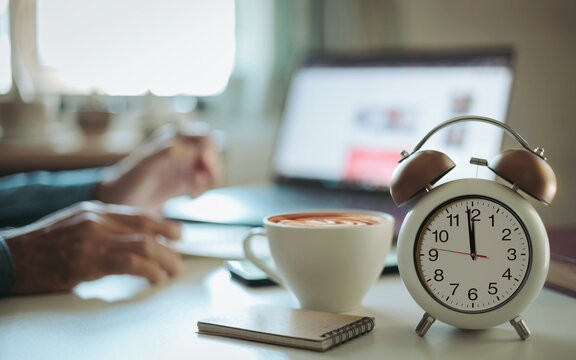 close up shot, focus alarm clock 12.00 PM. blurred businessman using laptop working and meeting online before breaking lunch