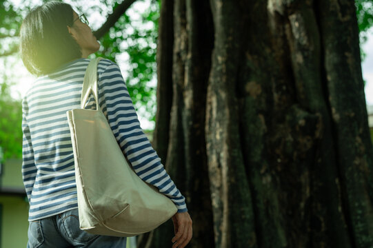 Asian woman carrying reusable canvas tote bag while admiring a tree in garden. Sustainable, eco friendly travel, mindfulness, nature connection and wellness. Responsible tourism, nature connection.