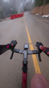 Vertical POV footage of a cyclist riding through a wet mountain road construction zone after rain, featuring safety barriers, winding roads and a tropical forest landscape.