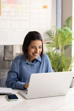 Smiling Latina businesswoman working online using laptop, reviewing information, reading good news in corporate email or message, enjoys successful digital workflow and positive business communication