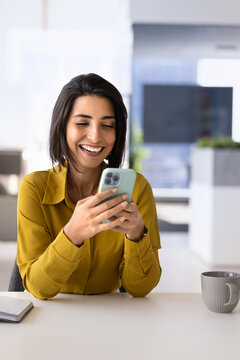 Businesswoman sits at office desk with smartphone, laughs, reading email, enjoy good business or work-related news in notice, take break during workday, check personal messages, chatting or calling