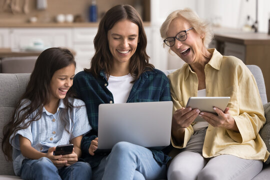 Three relatives women of diverse age sit on sofa with multiple digital gadgets. Older grandmother her young adult daughter and preteen granddaughter using laptop, phone and tablet. Fun, technologies