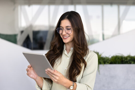 Smiling adult businesswoman standing in corporate office using digital tablet, working on project, checking information, browsing or replying on emails. Technology, professional application usage