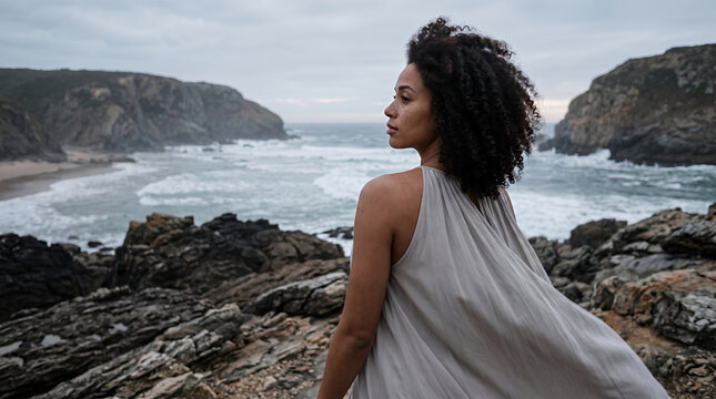 Woman in white sleeveless dress on rocky beach with dramatic ocean backdrop